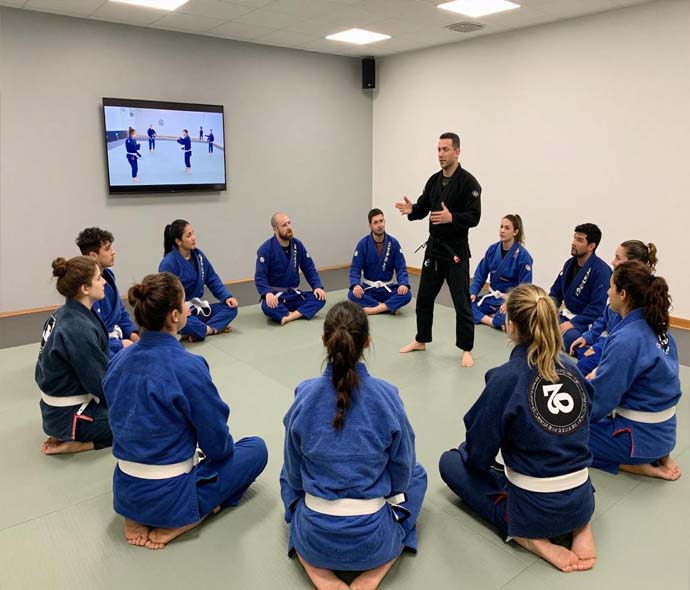 Instructor in black gi teaching a circle of students in blue BJJ gis with white belts on a gym mat