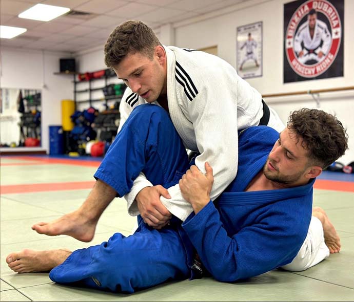 Two jiu-jitsu athletes in white and blue gis grappling on a gym mat during a sparring drill
