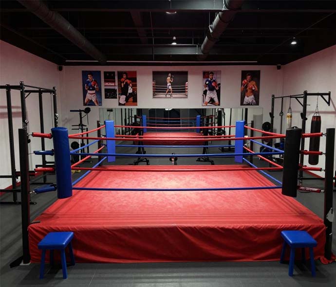 Red boxing ring with blue and red ropes and blue corner pads in a gym with mirrors, posters and heavy bags