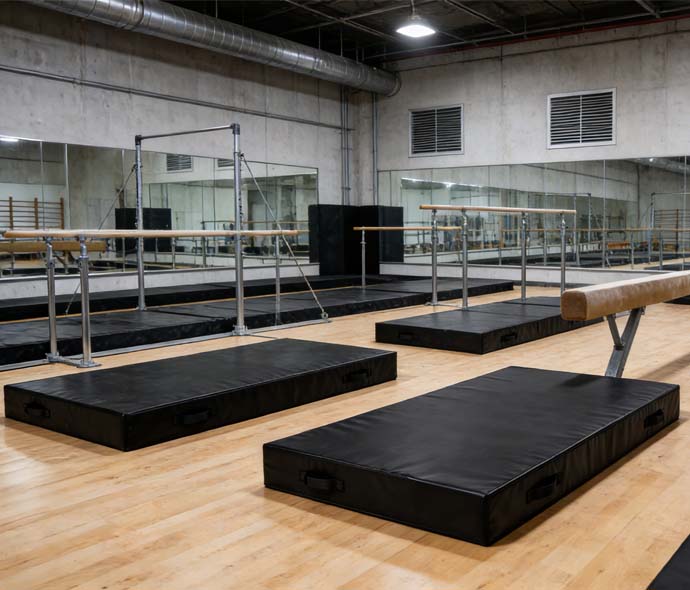 Black gymnastics crash mats with carry handles on a wood floor near parallel bars and a balance beam in a mirrored training gym