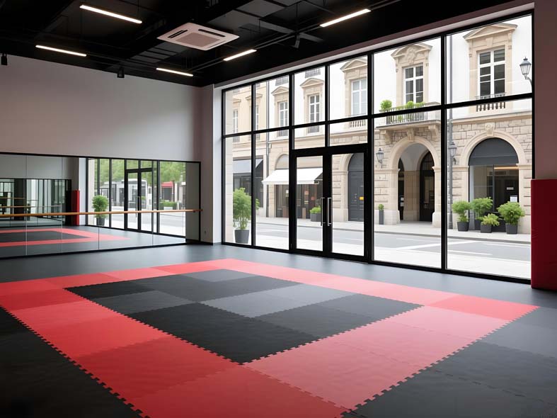 Red and black interlocking foam karate mats in a checkerboard layout on the floor of an empty dojo with large windows and mirrors