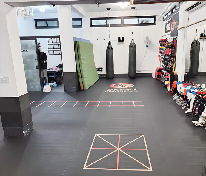 Black MMA training mat floor in a martial arts gym with agility ladder markings, heavy bags and gear rack
