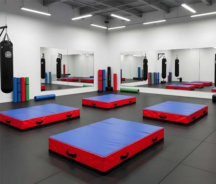 Four red and blue crash mats with carry handles on the dark rubber floor of a mirrored martial arts gym
