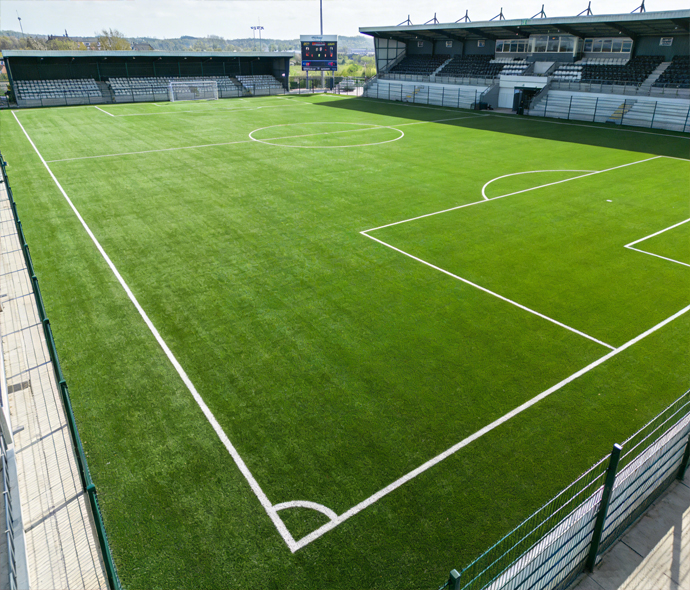 Artificial turf soccer field with white penalty area markings and a goal in front of covered stadium stands