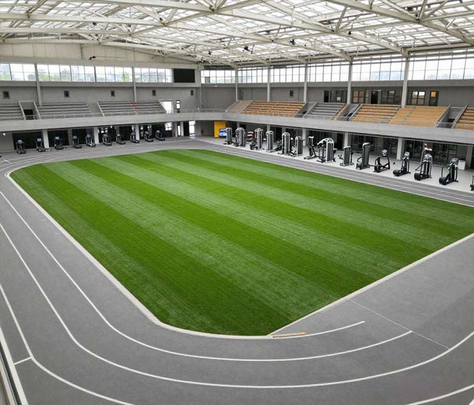 Indoor sports hall with a green artificial turf field surrounded by a grey running track under a steel roof
