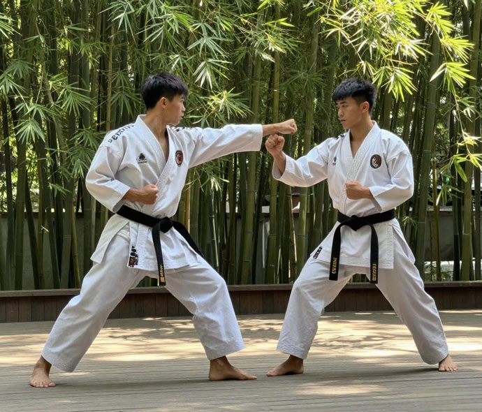 Two karateka in white uniforms and black belts sparring in a bamboo garden courtyard