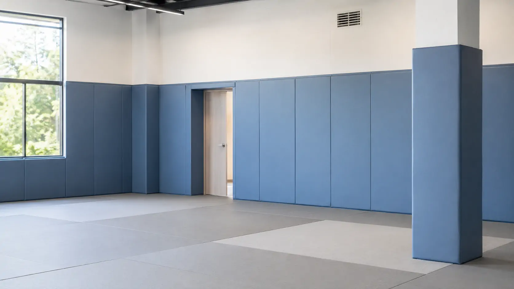 Blue padded wall panels and a wrapped column above gray floor mats in an empty gym room with a window and wooden door