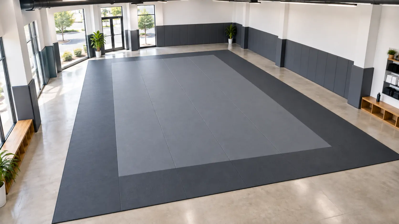 Large two-tone gray BJJ mat on polished concrete floor in an empty academy with wall padding and wooden cubby benches