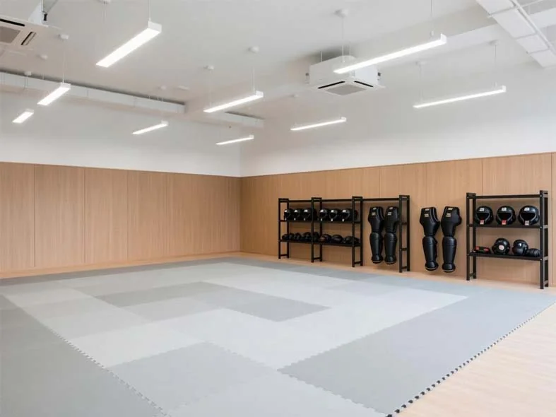 Gray interlocking foam karate mats covering a dojo floor beside racks of sparring helmets and shin guards