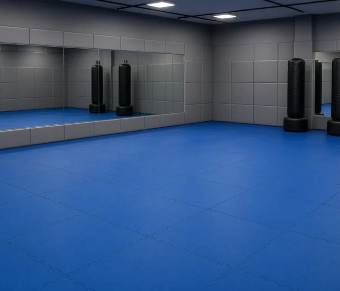 Blue interlocking MMA training mats covering a gym floor with gray padded walls, mirrors and freestanding punching bags